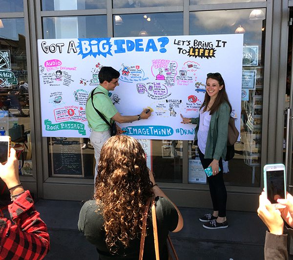 Visual strategist working on board depicting big ideas for DYBI book launch while crowd takes pictures.