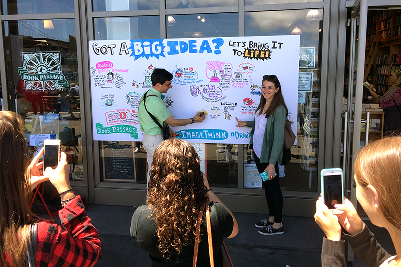 Visual strategist working on board depicting big ideas for DYBI book launch while crowd takes pictures.