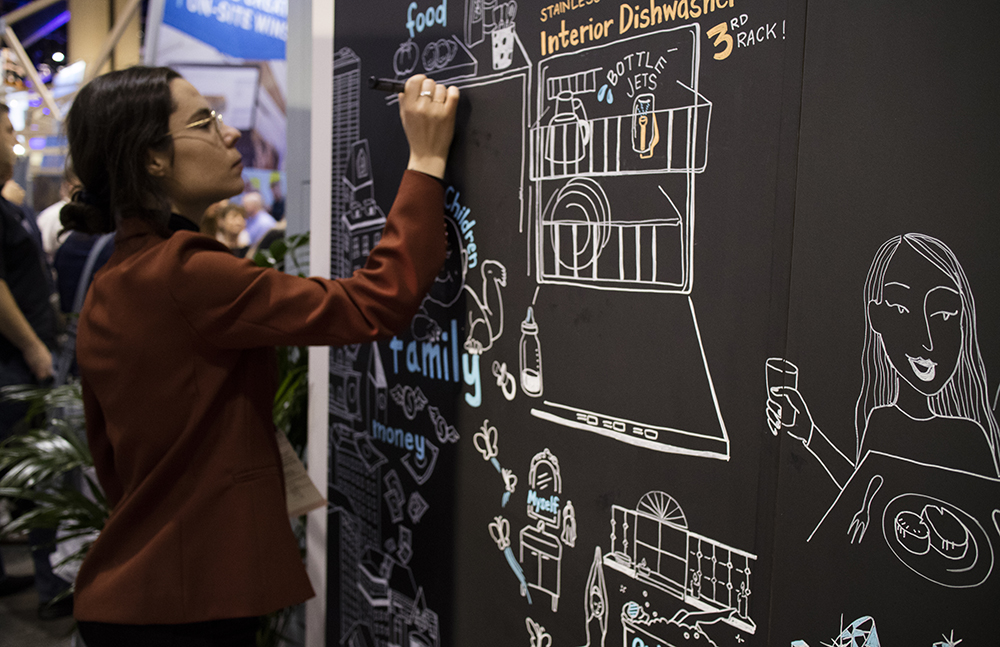 ImageThink graphic recorder Ona Rygelis scribes on a blackboard at a tradeshow booth.