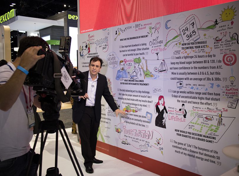 A camera man and reporter stand in front of an imagethink graphic recording at a healthcare tradeshow booth