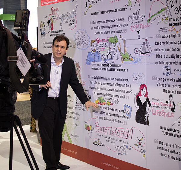 a camera man and reporter stand in front of an imagethink graphic recording at a healthcare tradeshow booth