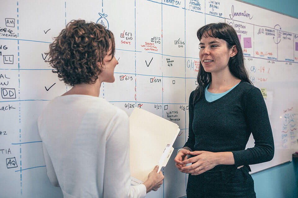 imagethink ceo nora herting and graphic recorder ona rygelis stand in front of a whiteboard calendar in their office
