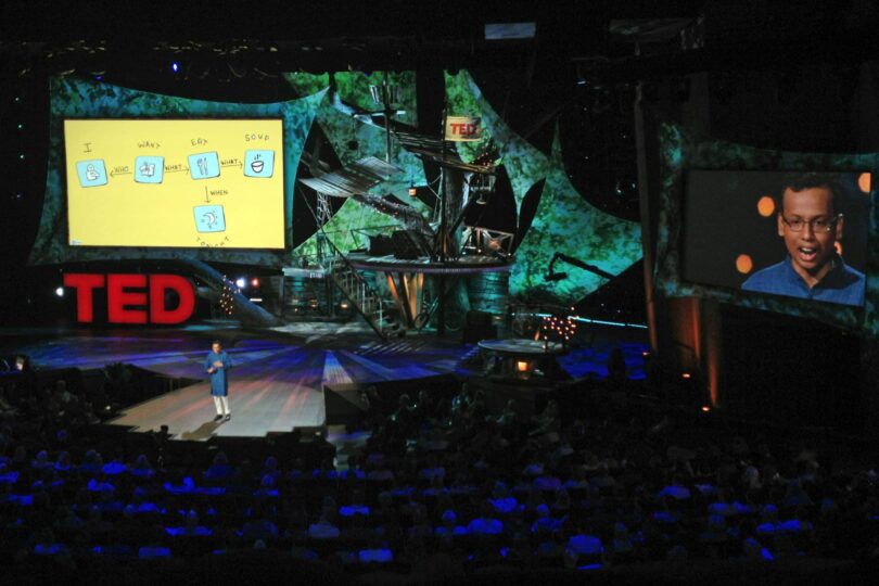 TED conference stage with a speaker presenting while ImageThink-style graphic recording and simple visual notes are projected on a large screen, turning a complex idea into a clear visual story for the audience.